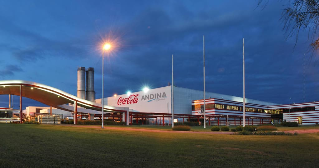 Fotografía profesional en toma alta de la sede corporativa de Coca-Cola Andina en un entorno urbano moderno. La escena está iluminada con luz dorada de atardecer, resaltando la fachada de vidrio y el branding icónico. La atmósfera es corporativa, de alta gama, y refleja liderazgo industrial confiable y progreso tecnológico.